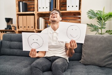 Canvas Print - Young hispanic man working on depression holding sad to happy emotion paper smiling and laughing hard out loud because funny crazy joke.