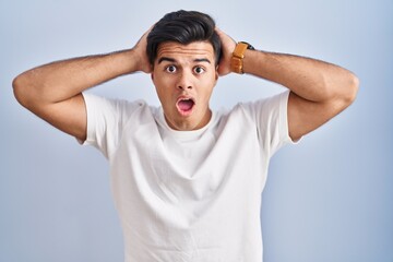 Wall Mural - Hispanic man standing over blue background crazy and scared with hands on head, afraid and surprised of shock with open mouth