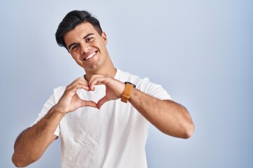 Poster - Hispanic man standing over blue background smiling in love doing heart symbol shape with hands. romantic concept.