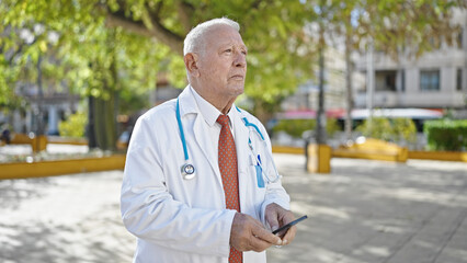 Wall Mural - Senior grey-haired man doctor standing with serious expression using smartphone at park