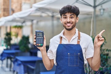 Wall Mural - Arab man with beard wearing waiter apron at restaurant terrace holding dataphone smiling happy pointing with hand and finger to the side