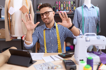 Hispanic man with beard dressmaker designer working at atelier showing and pointing up with fingers number nine while smiling confident and happy.