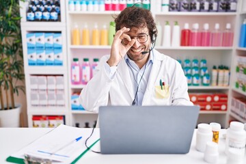 Wall Mural - Hispanic young man working at pharmacy drugstore working with laptop smiling happy doing ok sign with hand on eye looking through fingers