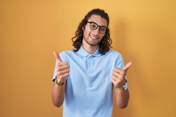 Poster - Young hispanic man standing over yellow background success sign doing positive gesture with hand, thumbs up smiling and happy. cheerful expression and winner gesture.