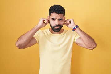 Poster - Hispanic man with beard standing over yellow background covering ears with fingers with annoyed expression for the noise of loud music. deaf concept.