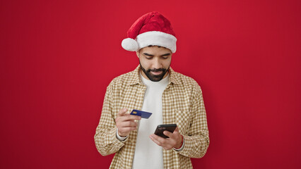 Sticker - Young hispanic man shopping with smartphone and credit card wearing christmas hat over isolated red background