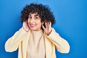 Poster - Young brunette woman with curly hair standing over blue background trying to hear both hands on ear gesture, curious for gossip. hearing problem, deaf