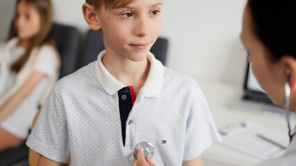 Wall Mural - Doctor and kid patient are in the clinic. Physician in white coat examining a serious young boy with a stethoscope, close up. Medicine, therapy concept