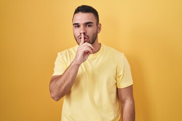 Sticker - Young hispanic man standing over yellow background asking to be quiet with finger on lips. silence and secret concept.