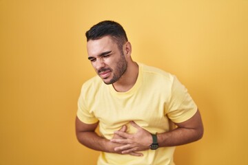 Canvas Print - Young hispanic man standing over yellow background with hand on stomach because indigestion, painful illness feeling unwell. ache concept.