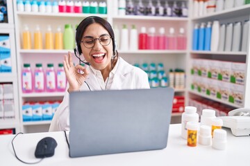 Wall Mural - Young arab woman working at pharmacy drugstore using laptop smiling positive doing ok sign with hand and fingers. successful expression.