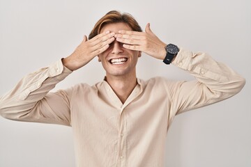 Wall Mural - Young man standing over isolated background covering eyes with hands smiling cheerful and funny. blind concept.