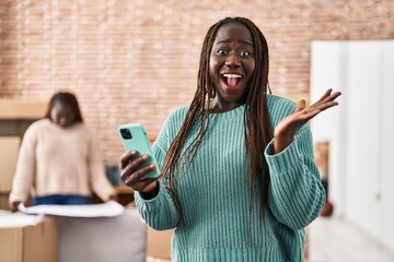 Sticker - African woman using smartphone at new home celebrating victory with happy smile and winner expression with raised hands