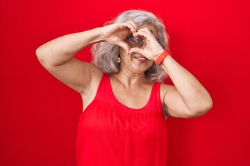 Wall Mural - Middle age woman with grey hair standing over red background doing heart shape with hand and fingers smiling looking through sign