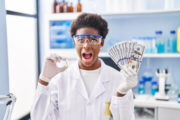 Canvas Print - African american woman working at scientist laboratory holding dollars angry and mad screaming frustrated and furious, shouting with anger looking up.