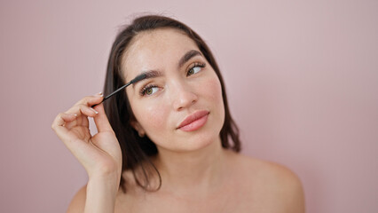 Young beautiful hispanic woman applying make up on eyebrows over isolated pink background