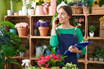 Wall Mural - Young beautiful hispanic woman florist talking on smartphone reading document at flower shop