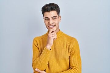 Sticker - Young hispanic man standing over blue background looking confident at the camera smiling with crossed arms and hand raised on chin. thinking positive.