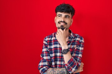 Wall Mural - Young hispanic man with beard standing over red background with hand on chin thinking about question, pensive expression. smiling and thoughtful face. doubt concept.