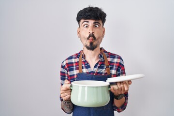 Canvas Print - Young hispanic man with beard wearing apron holding cooking pot making fish face with mouth and squinting eyes, crazy and comical.