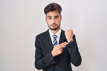 Poster - Young hispanic man with tattoos wearing business suit and tie in hurry pointing to watch time, impatience, looking at the camera with relaxed expression