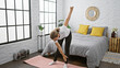 Serious young blonde woman finding balance, strengthening her concentration with morning yoga exercise on her bed, warming up in cozy indoor bedroom space, enjoying calmness and relaxation.