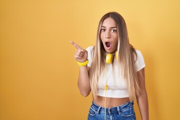 Wall Mural - Young blonde woman standing over yellow background wearing headphones surprised pointing with finger to the side, open mouth amazed expression.