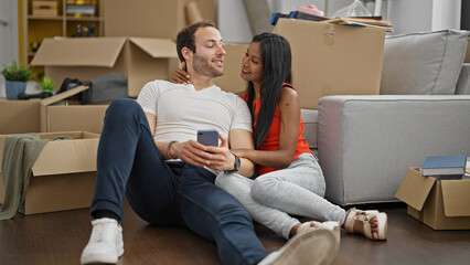 Wall Mural - Beautiful couple sitting on floor together using smartphone smiling at new home