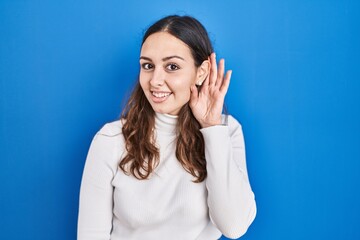 Sticker - Young hispanic woman standing over blue background smiling with hand over ear listening an hearing to rumor or gossip. deafness concept.