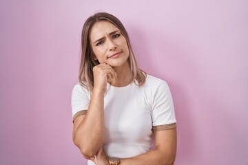 Poster - Blonde caucasian woman standing over pink background with hand on chin thinking about question, pensive expression. smiling and thoughtful face. doubt concept.