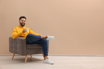 Sticker - Handsome man with cup of drink sitting in armchair near beige wall indoors, space for text