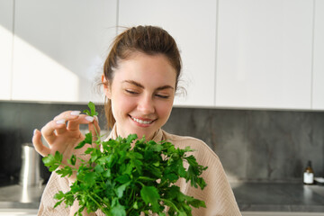 Portrait of attractive girl with bunch of parsley, eating fresh herbs and vegetables, cooking in the kitchen