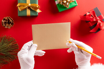 Poster - Santa claus holding envelope and pencil with copy space and christmas presents on red background