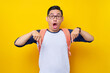 © Bangun Stock Photo - Shocked young Asian student man in casual clothes and glasses backpack pointing finger down isolated on yellow background. Education in High School University College concept