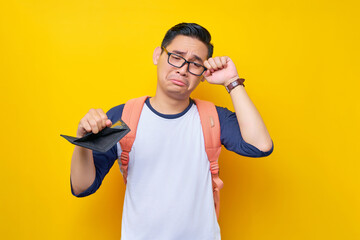 Problem Financial. Sadness young Asian student man in casual clothes and glasses backpack showing an empty wallet isolated on yellow background. Education in High School University College concept