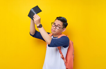 Wall Mural - Problem Financial. Young Asian student man in casual clothes and glasses backpack looking at an empty wallet isolated on yellow background. Education in High School University College concept