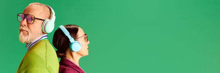 Senior man and woman standing back to back, listening to music in headphones against green studio background. Concept of beauty and fashion, relationship, modern style, age. Banner