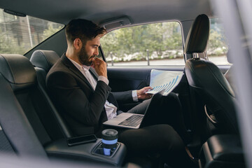 Wall Mural - Male entrepreneur sitting on car backseat and working with documents while going to office