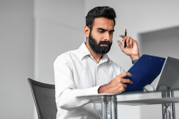 Serious indian bearded man sitting at table with laptop and clipboard, working with documents in office interior