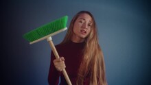 Portrait Of Young Beautiful Caucasian Girl With Long Brown Hair In Burgundy Sweater Looking At The Camera And Waving Cleaning Green Brush On Blue Background. Protest, Fight For Female Rights, Freedom