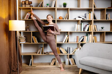 Young Woman In Sportswear Stretching Legs, Doing Splits Standing In Library And Reading Book. Female In Daily Workout Make Yoga Training At Home