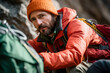 Bearded climber in an orange jacket setting up camp in the mountains at a rest stop.