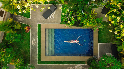 Bird's eye view of a blue water swimming pool located in the jungle on a tropical. A young beautiful woman in a red swimsuit lies in the pool. Vacation and travel concept to tropical islands.