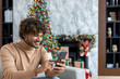 © Liubomir - Young handsome hispanic man at home sitting on sofa in living room for Christmas near tree, man using phone happily smiling typing online text message and browsing internet pages.