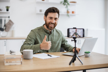 Wall Mural - Confident bearded man sitting at desk and working on wireless laptop while recording video on modern smartphone fixed on tripod showing thumb up