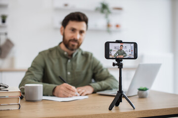 Wall Mural - Happy bearded guy using smartphone and tripod for creating new video content in social media. Positive male blogger in casual attire having interaction with subscribers during online communication.