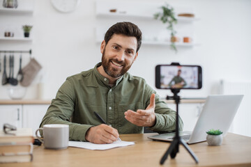 Wall Mural - Happy bearded guy using smartphone and tripod for creating new video content in social media. Positive male blogger in casual attire having interaction with subscribers during online communication.