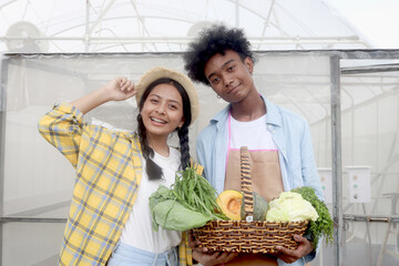 Wall Mural - Portrait of two multiethnic teenager friend, Asian girl and African boy holding fresh harvest fruit vegetable basket at front of vegetable farm greenhouse, young gardener working in agricultural field