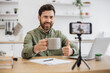 © sofiko14 - Happy attractive streamer sitting at table near modern laptop and recording video content on smartphone fixed on tripod showing thumb up