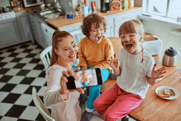 Young mother taking a selfie with her children on a smartphone while eating breakfast and being messy in the kitchen